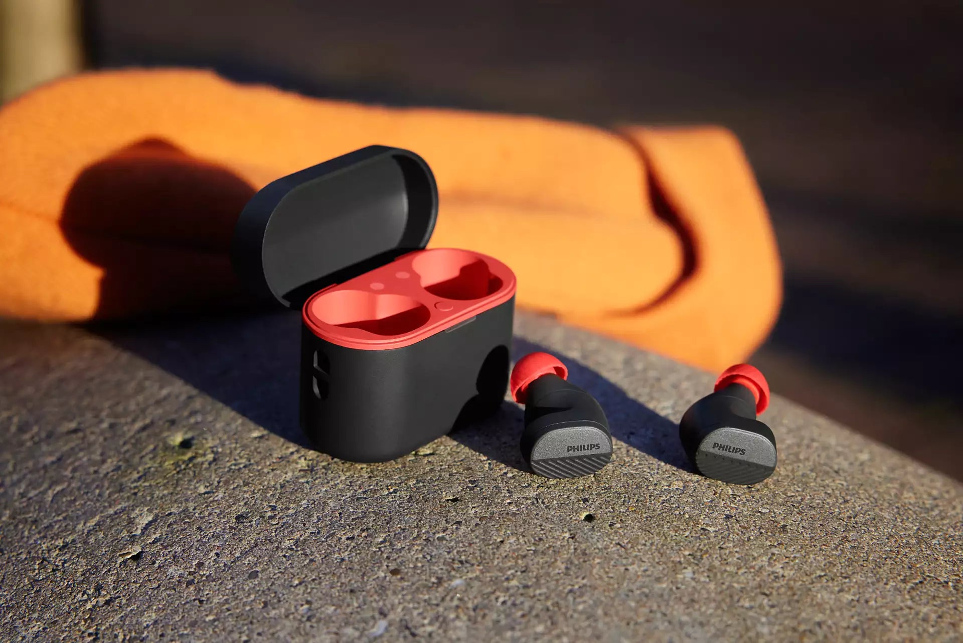 Ægte trådløse hovedtelefoner fra Philips model TAA5508BK/00 med en sort og rød opladningsæske liggende på en grå overflade skabt af beton headset med runde ørepropper placeret ved siden af æsken og et orange stof synligt i baggrunden