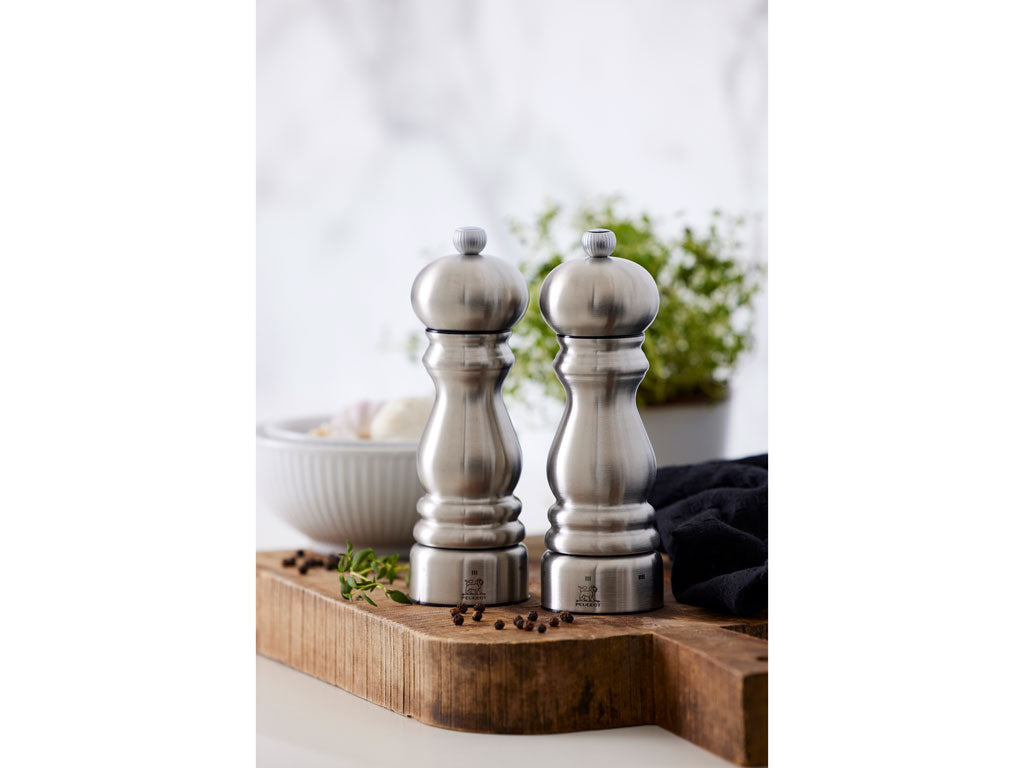 Producent Peugeot Paris peberkværn i stål med en højde på 18 cm står på et skærebræt i træ sammen med krydderier og en skål med ingredienser bagved. Kværnen har en elegant form med en stor knop på toppen og giver et moderne udtryk til køkkenet.