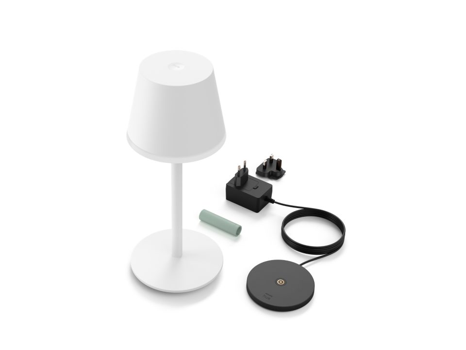 Billede af Philips Hue Go bærbar bordlampe i hvid farve placeret sammen med en opladeradapter, et kabel og en grøn batteriscylinder. Lampen har en moderne design med en rund base og en kegleformet skærm. Opladeren har forskellige stik til strøm og et rundt dock til tilslutning.