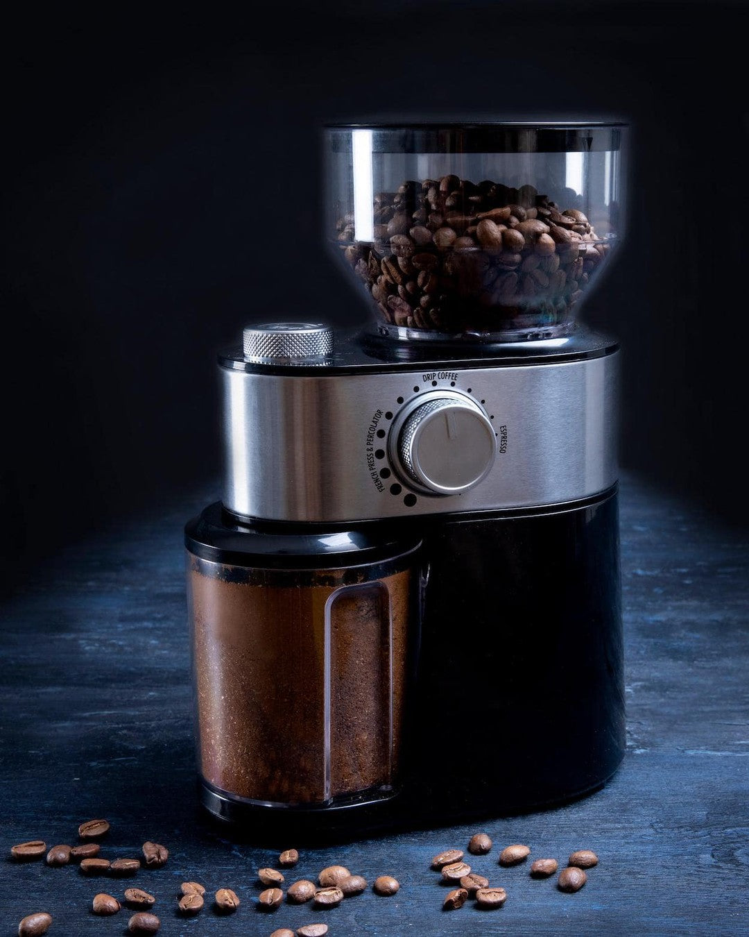 Gastronoma kaffekværn i sort og rustfrit stål med 200W motor som har en glaskapacitet til hele kaffebønner og en beholder til det malede kaffe med kaffebønner spredt omkring den på en mørk overflade