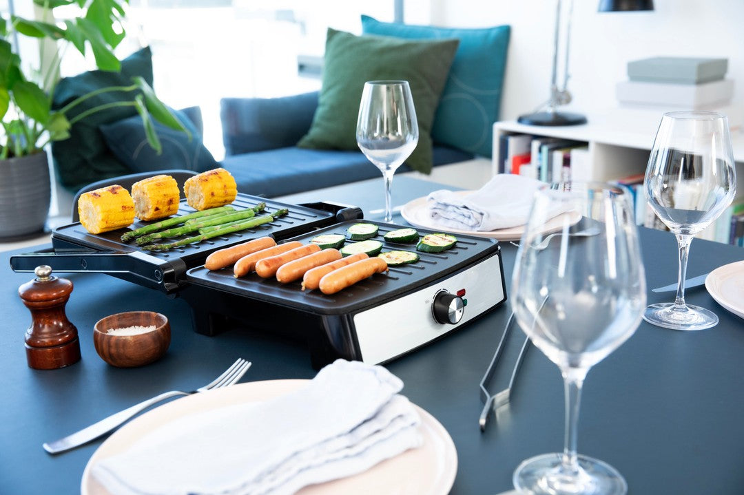 Et bord dækket til middag med Gastronoma Panini grill i sort og stål med 2200W. Grillen har hotdogs, zucchini, asparges og grillede majskolber. Der er glas og tallerkener klar til servering, samt en peberkværn og en skål med salt på bordet. Baggrunden viser en komfortabel stue med farverige puder.