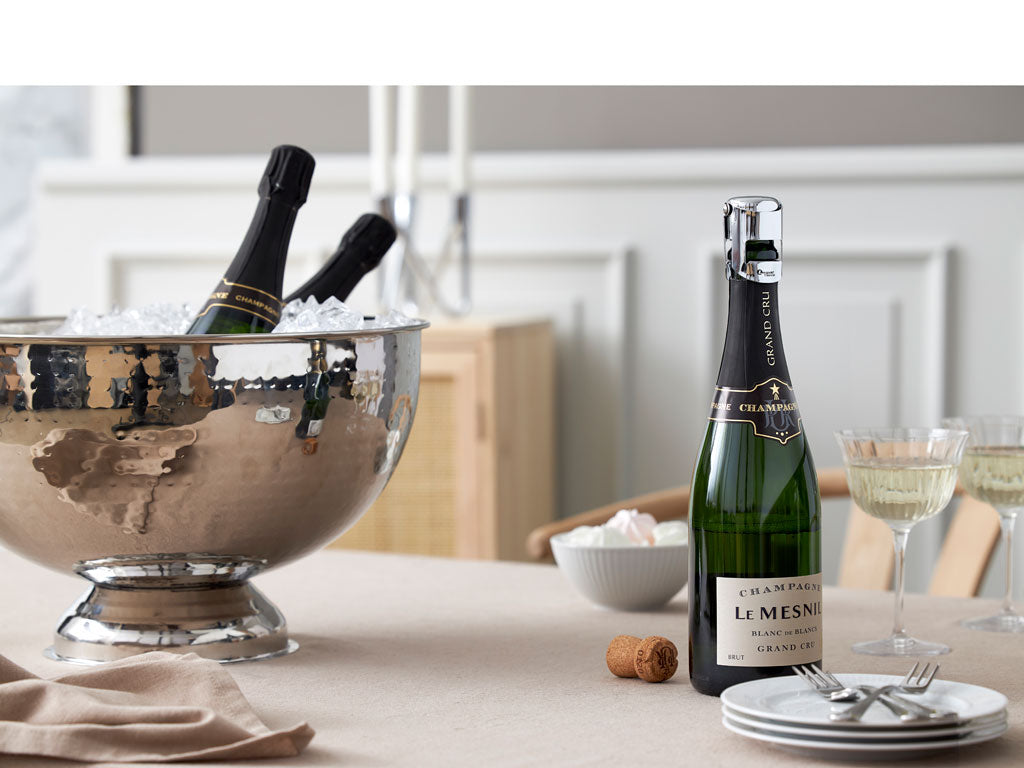 Champagnebowle i hamret blank stål med dets spejlfornemmelse fyldt med is og champagneflasker samt et sæt glas og en skål med dessert på et dækket bord