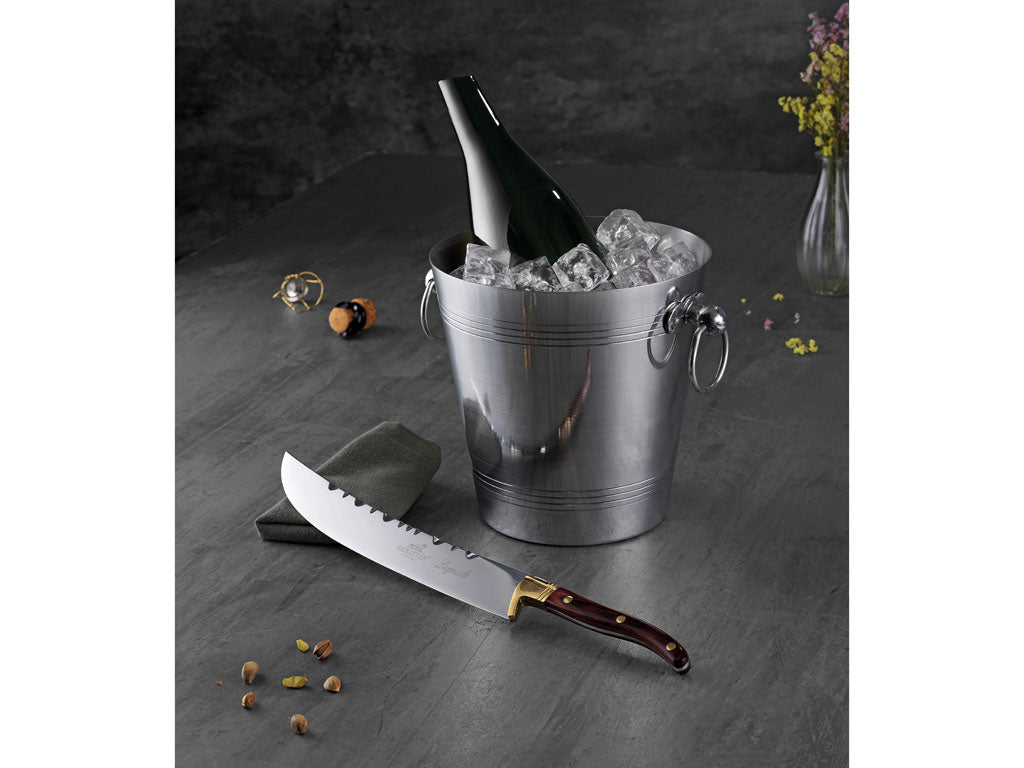 Aluminium champagnekøler med is og en flaske champagne stående i køleren ved siden af en elegant kniv med træskaft på en grå bordplade