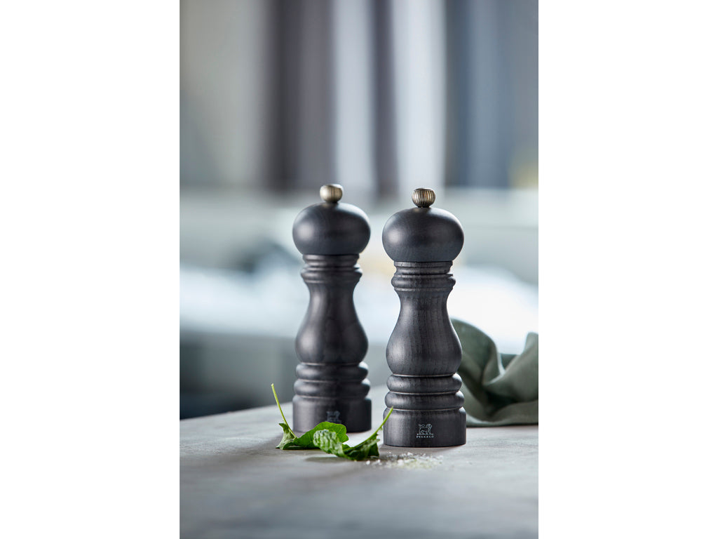 Peugeot Paris salt- og peberkværnsæt i grafit farve med en højde på 18 cm placeret på en bordplade med grønne krydderurter i forgrunden og en neutral baggrund.