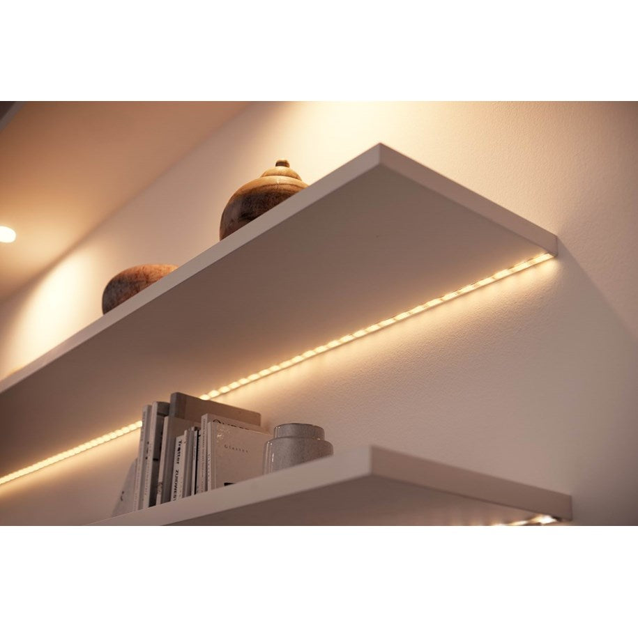 LED strip med varmt lys monteret under en hvid hylde, der viser bøger og dekorative genstande.