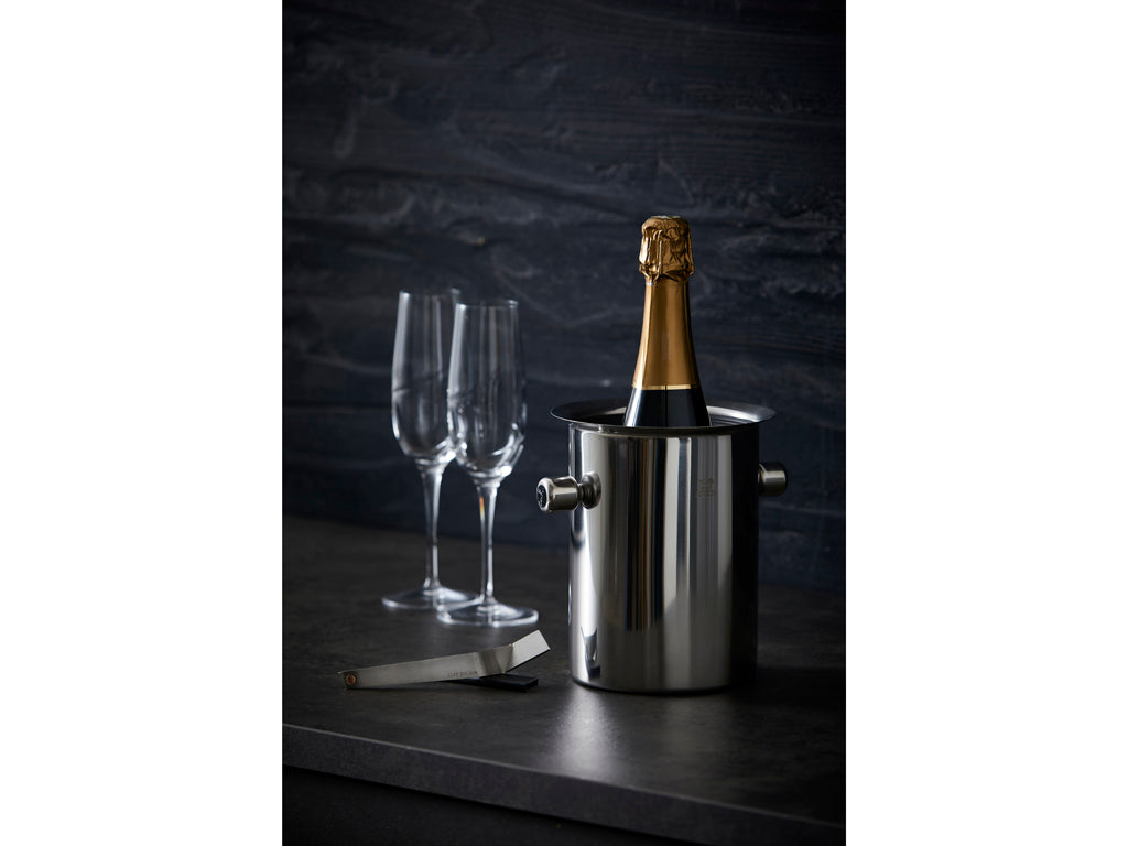 Peugeot champagnekøler i rustfrit stål med en flaske champagne indsat, to elegante glas ved siden af og en kapselkniv på bordet.
