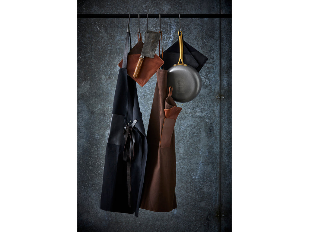 Pillivuyt Gourmet Forklæde i sort hængende sammen med forskellige køkkenredskaber på en metalstang mod en grå væg