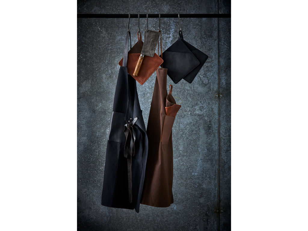 Billede af Pillivuyt Gourmet Grydelap i størrelse 20,5 x 20,5 cm i brun farve hængende sammen med andre køkkenredskaber på en metalstang mod en mørk baggrund