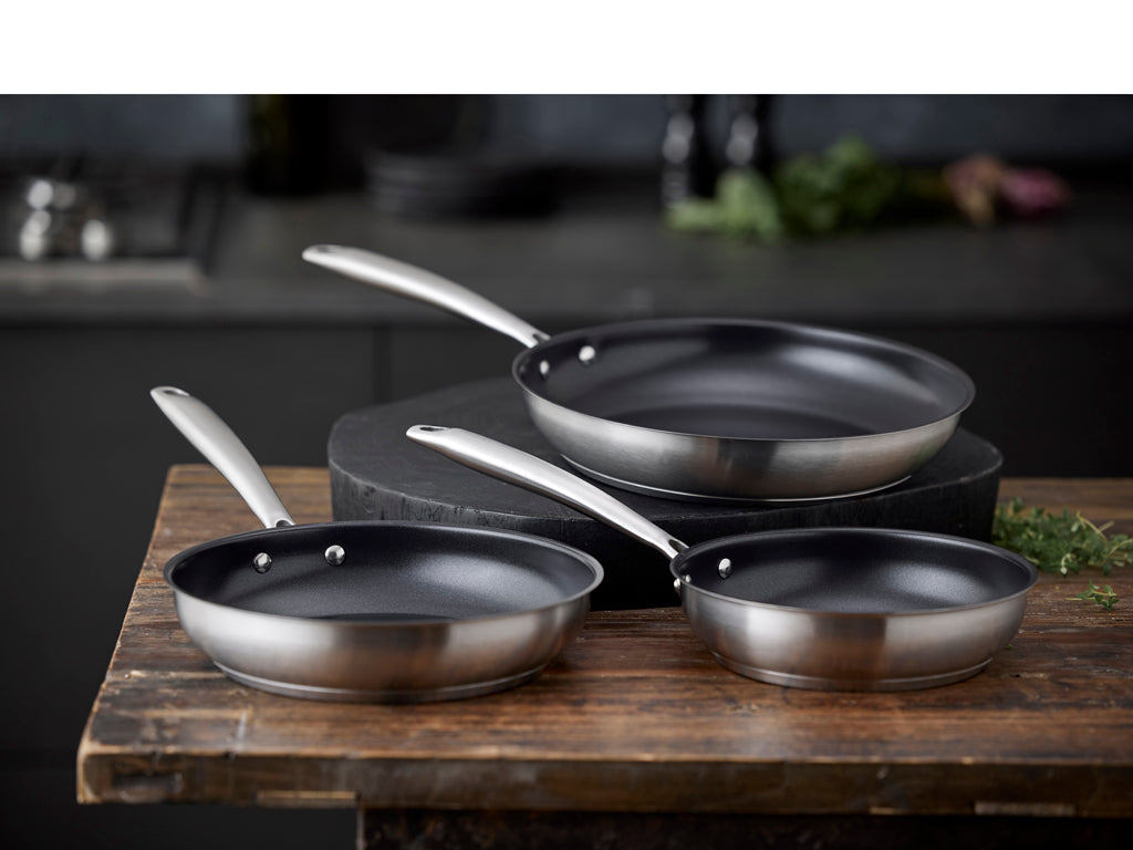 Tre keramiske non-stick stegepander fra Pillivuyt Gourmet Roya med diameter på 28 cm i stål placeret på et træbord i et moderne køkkenmiljø.