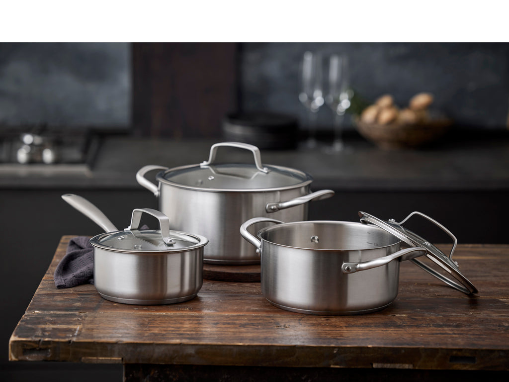 Design af tre gryder i rustfrit stål fra Pillivuyt Gourmet med glaslåg placeret på en træoverflade, hvor den mindste gryde har en håndtag og den største gryde står delvist åben, og to elegant glas i baggrunden.