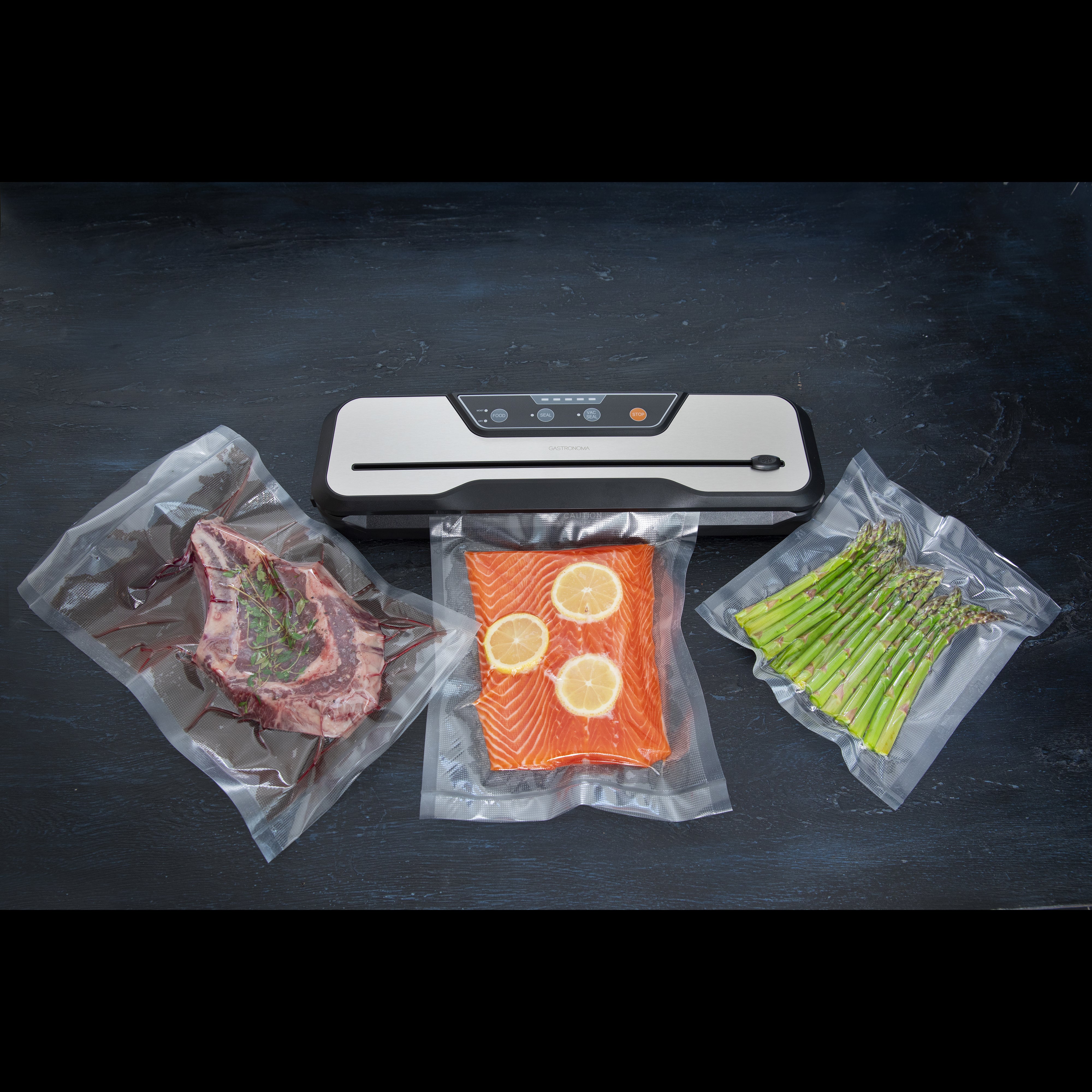 Gastronoma vakuumpakker i sort og rustfrit stål med 110W placeret centralt med pakker af vakuumforseglet kød, fisk med citron skiver og asparges omkring den
