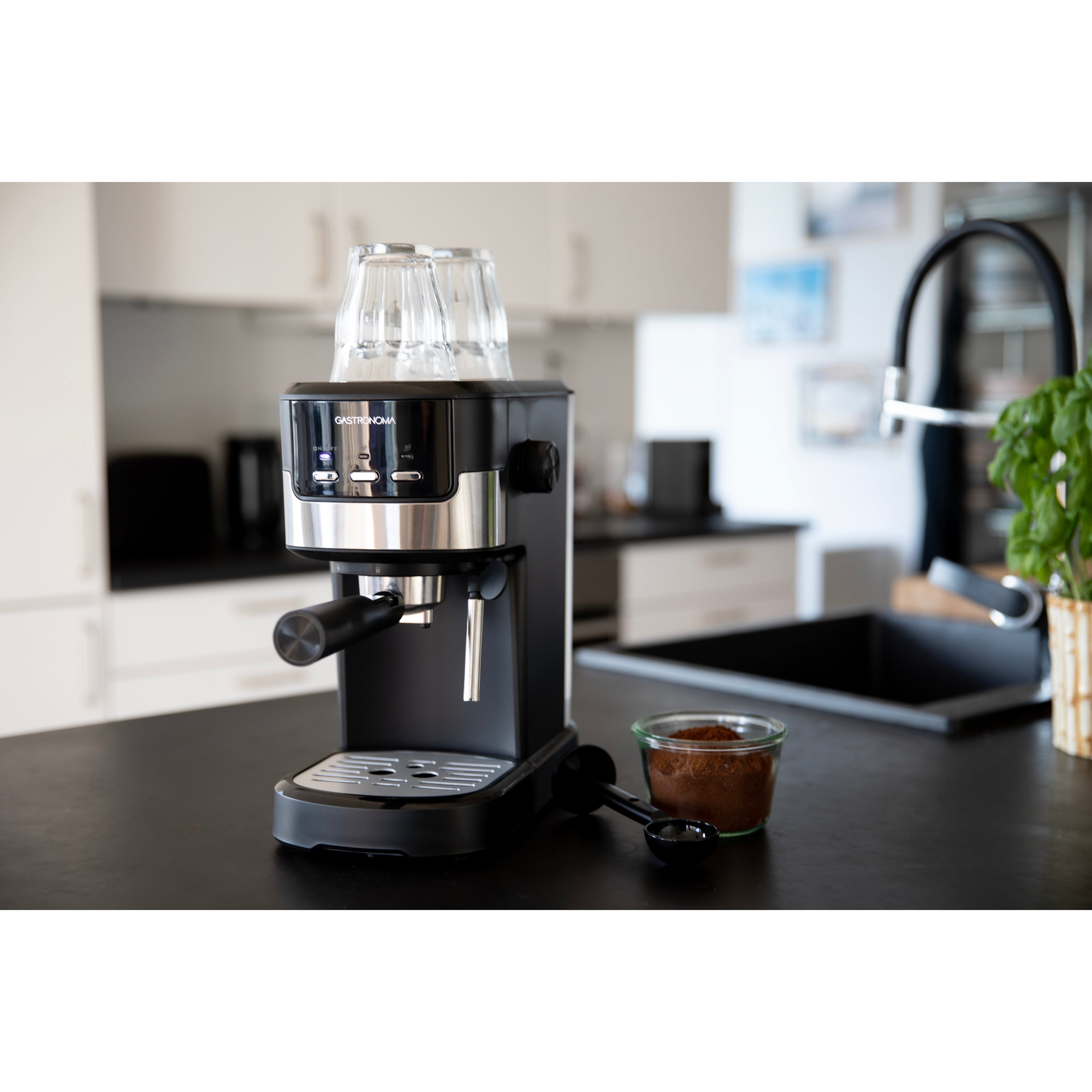 Gastronoma espressomaskine med 15 bar tryk og 1100W effekt står på et køkkenbord med et moderne design strålende med metal og sort finish glasbehållere placeret på toppen samt en skefuld kaffe ved siden af en glasbeholder fyldt med malet kaffe