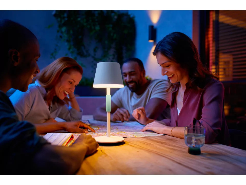 En bærbar bordlampe fra Philips Hue Go i hvid farve lyser op på et bord, hvor fire mennesker samles for at se på et kort i en hyggelig aftenstemning med blødt lys og afslappet atmosfære