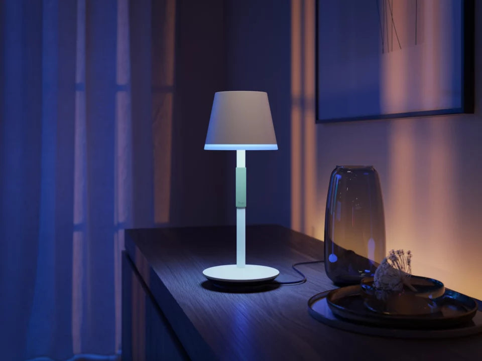 Philips Hue Go bærbar bordlampe i hvid farve med en klassisk skærm og en moderne design placeret på et træbord omgivet af blødt lys og interiørdekorationer