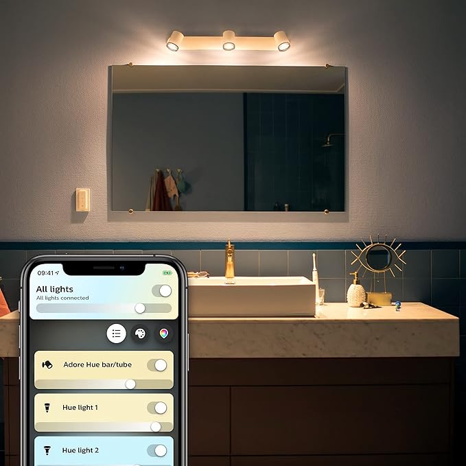 Billede af et badeværelse med Philips Hue Adore White Ambiance spotlys monteret over et spejl der reflekterer lysene et moderne badeværelsesmøbel en smartphone viser et smart home interface til styring af lysene og en håndvask med kosmetikartikler og en dekorativ genstand