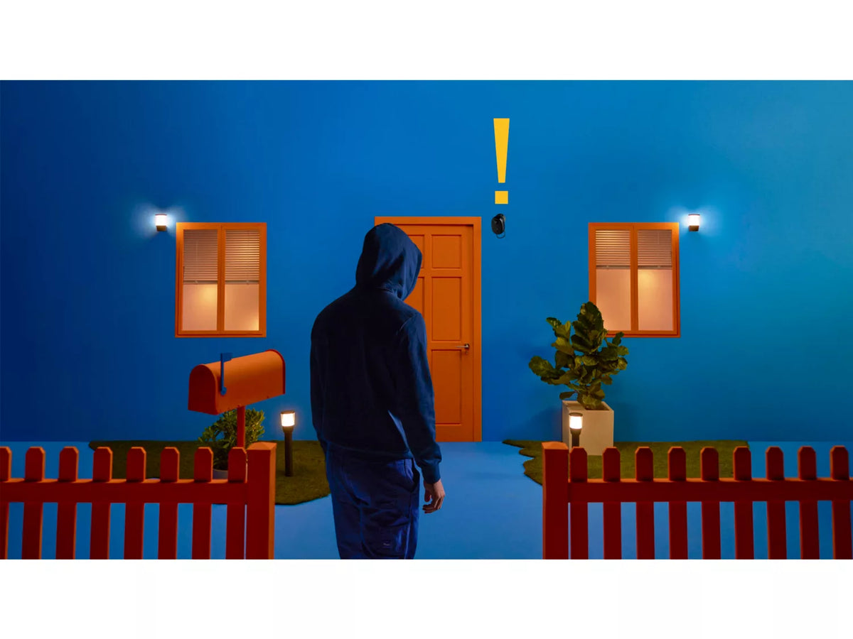 Person iført hætte trækker op til en farverig blå bygning med orange dør og vinduer kamera er placeret over døren der er en postkasse og belysning i haven med en grøn plante i juletiden en tilstedeværelse med et advarselssymbol viser en potentiel sikkerhedssituation