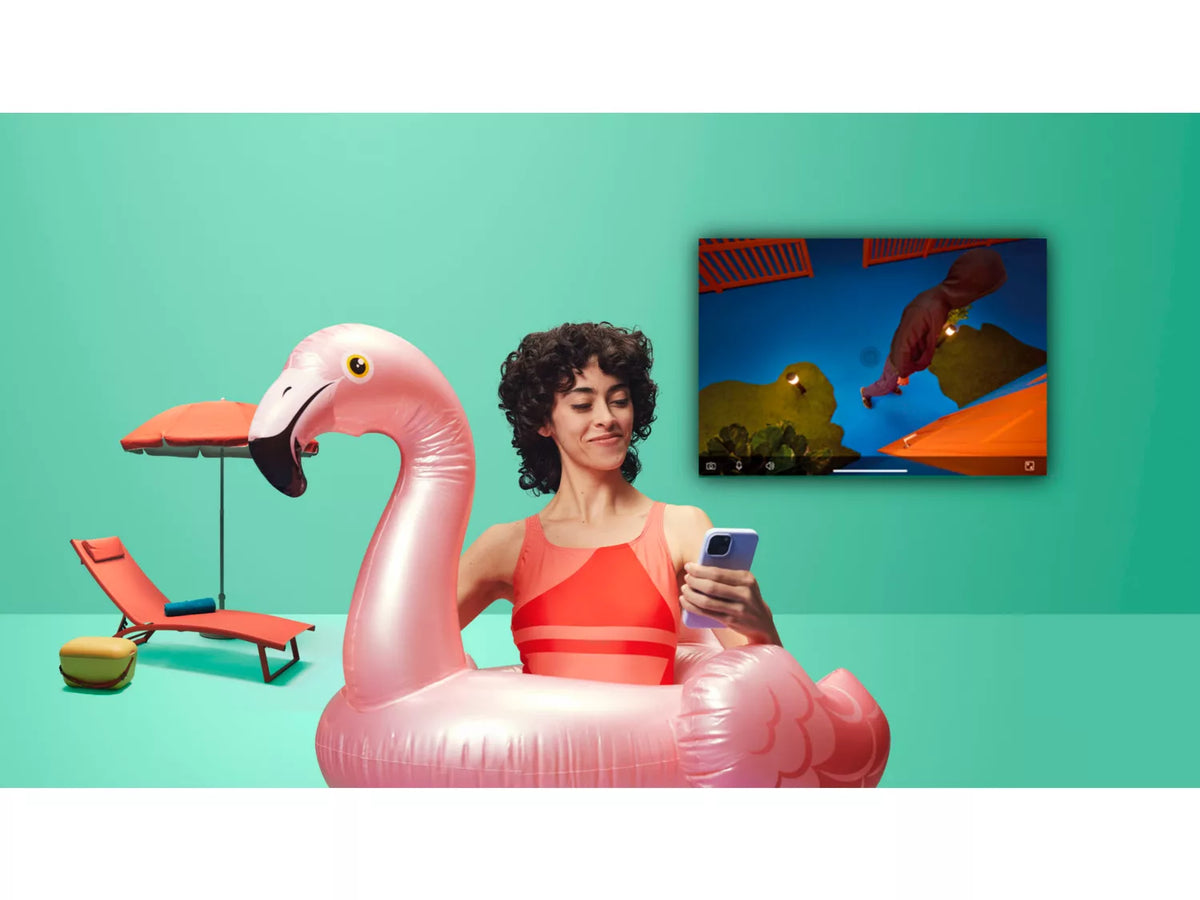 En kvinde sidder i en oppustelig flamingo i en pool, hun holder en smartphone og ser på en TV-skærm i baggrunden med en visning af udendørs aktiviteter, derudover er der en sammenklappelig solseng og parasol i nærheden.