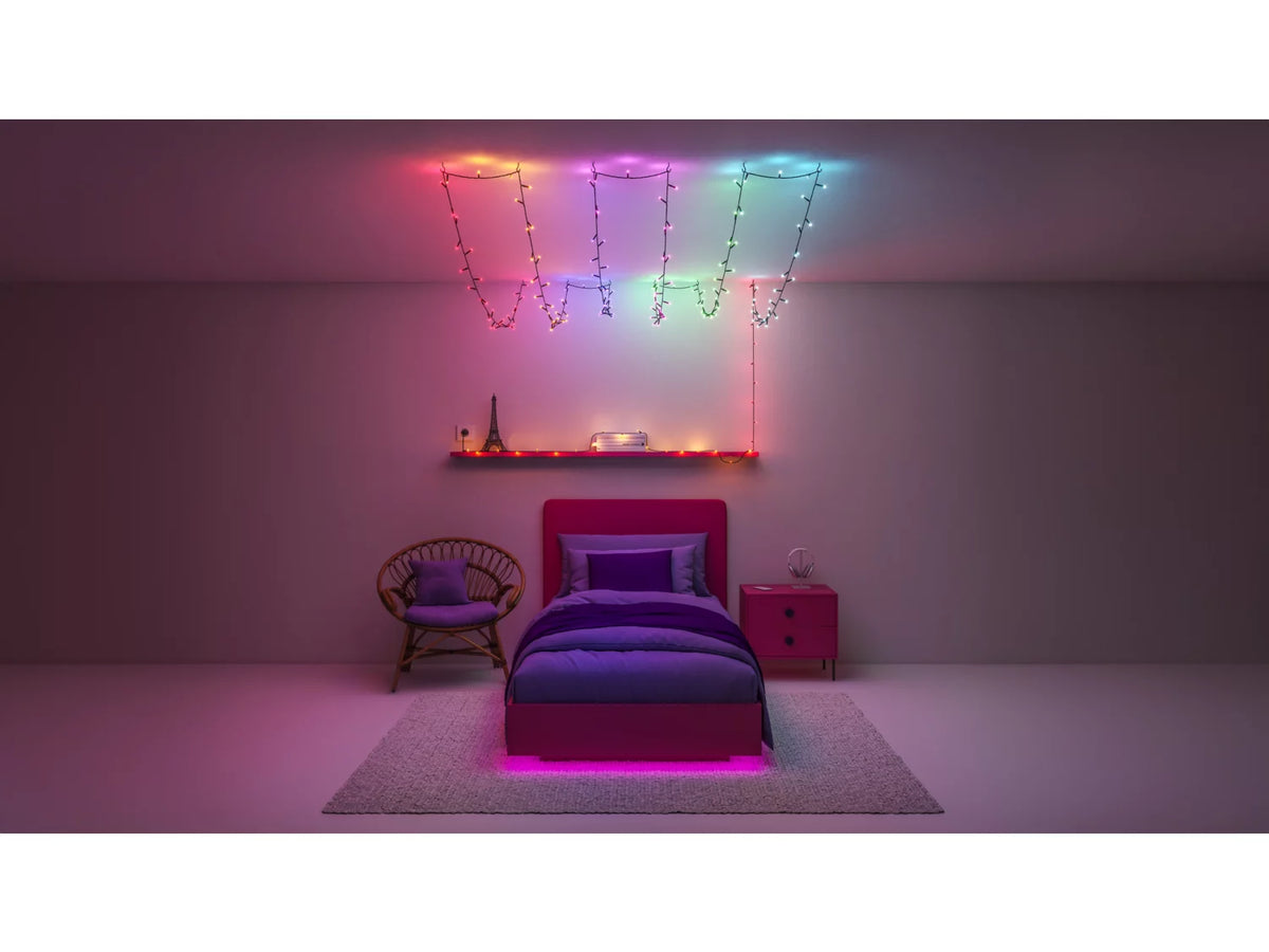 Et værelse med WIZ Fairy light lyskæde på 20 meter der hænger fra loftet i farverne pink, blå og grøn. Der er en seng med lilla sengetøj og en lilla sengegavl. En minimalistisk lampe står på en hylde og en stol i rattan er placeret i hjørnet. Gulvet er dækket med et blødt, lyst tæppe.