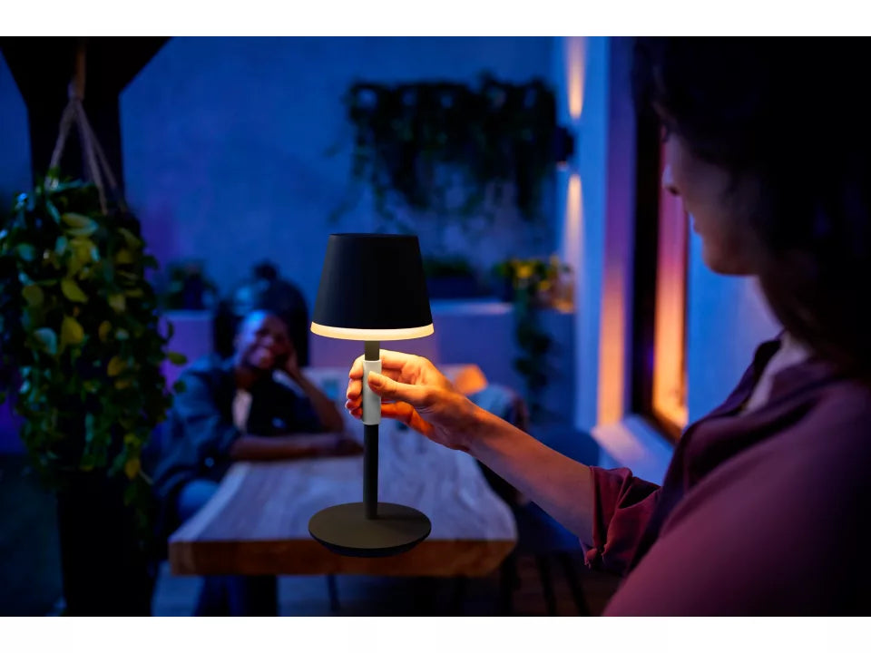 Billede af en person, der holder en sort Philips Hue Go bærbar bordlampe med en rund fod og en lysende lampehjelm i et moderne udendørs miljø om aftenen med blød belysning og grønne planter i baggrunden.