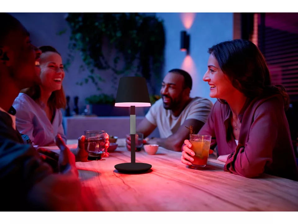 Billede af en gruppe mennesker, der sidder omkring et bord og griner, med en Philips Hue Go bærbar bordlampe i sort, der lyser i skiftende farver på bordet i en hyggelig atmosfære om aftenen.