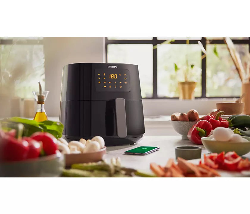 Philips HD9280/90 Connected Airfryer XL i sort farve står på et køkkenbord omgivet af friske grøntsager og madlavningsredskaber med en smartphone i nærheden og et skilt med temperaturen sat til 180 grader