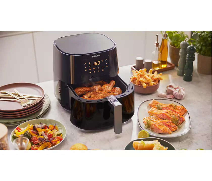 Billede af Philips HD9280/90 Connected Airfryer XL som frituresteger kyllingevinger og laks med digitalt kontrolpanel der viser temperaturen 180 grader omgivet af forskellige retter som salat og pommes frites på et køkkenbord