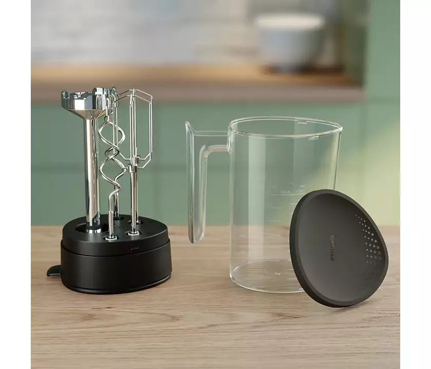 Philips HR3781/10 kraftig håndmixer med en effekt på 500 W står på en træoverflade sammen med en gennemsigtig målebæger og et sort låg ved siden af den centrale enhed som har piskeris og dejkrog