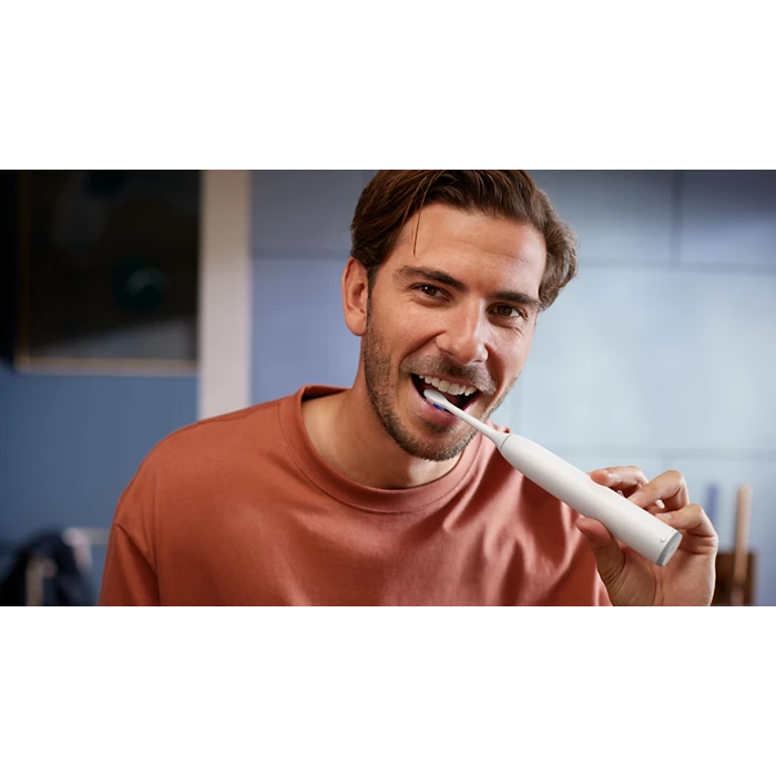 Mand med et smil børster tænder med en Philips Sonicare 6500 genopladelig tandbørste HX7410/02 i et moderne køkkenmiljø.