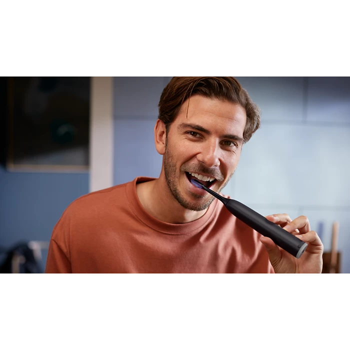 Mand med mørkt hår bruger en Philips Genopladelig Eltandbørste Sonicare Serie 7100 HX7421/01 mens han smiler med børsten i munden i et moderne køkkenmiljø.