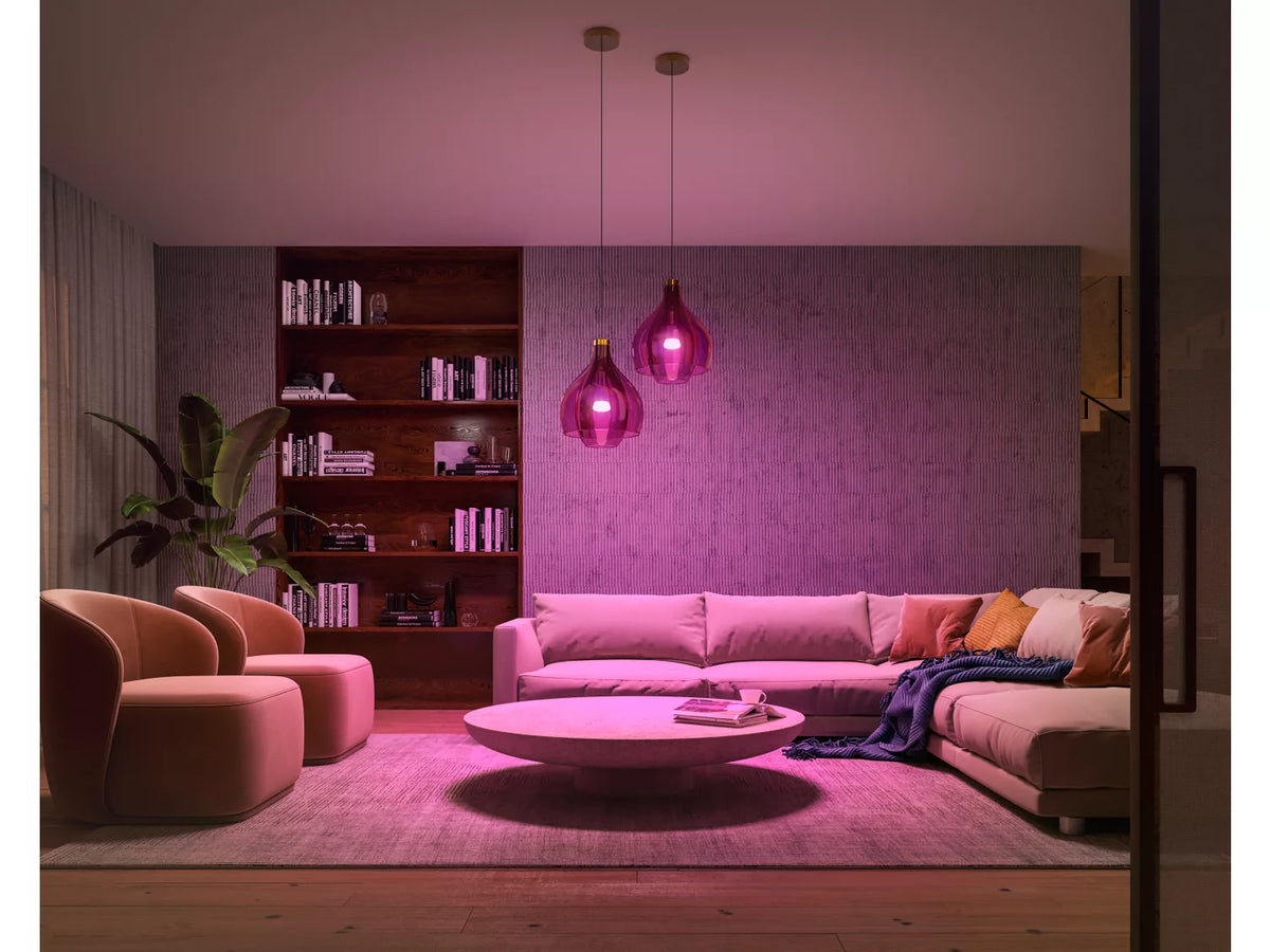 Stue med moderne indretning oplyst af Philips Hue E27 pærer i en varm pink farve to hængende lamper over en hvid sofa med puder og et runde bord i midten der står på et blødt tæppe og et bogreol i baggrunden
