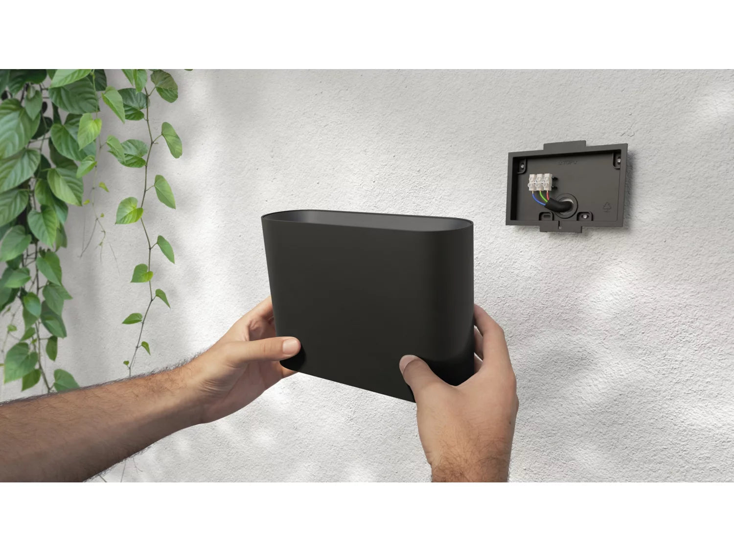 Hånd holder en sort Philips Hue Dymera væglampe klar til montering på en hvid væg med planter omkring det monteringsbeslag, som viser ledningerne i baggrunden