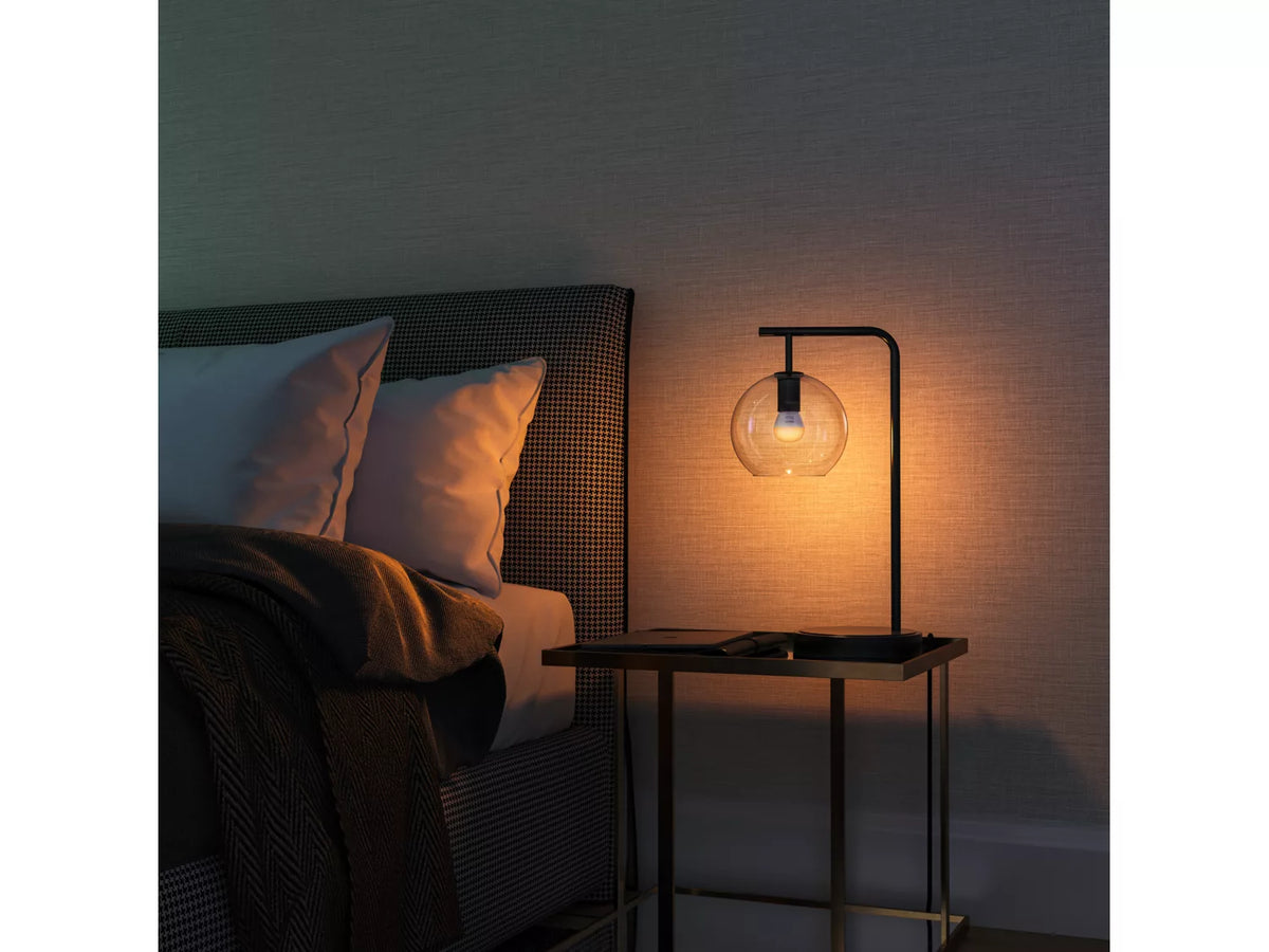 Philips Hue White Ambiance E14 pærer i en moderne lampe ved siden af en seng med puder og tæpper i en dæmpet atmosfære
