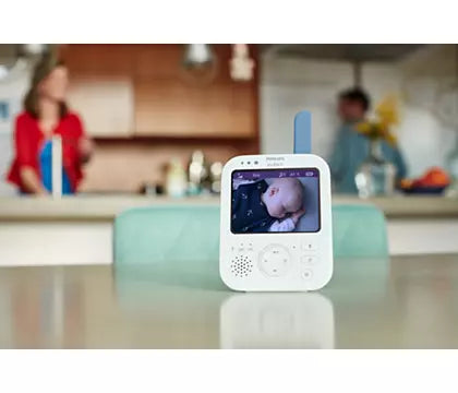 Digital babyalarm Philips Avent SCD845/26 med video står på et bord i køkkenet med et billede af en baby på skærmen og forældre i baggrunden