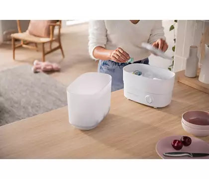 En person ved et træbord rengør en Philips Avent SCF291/00 sterilisator til sutteflasker mens der er en uåbnet sterilisator ved siden af samt skåle med frugt på bordet og en stol i baggrunden