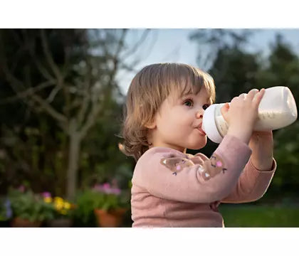 Barn drikker fra en Philips Avent Natural Response sutteflaske udendørs i sollys med grønne planter og blomster i baggrunden. Sutteflasken er i hånden på et barn, der ser tilfreds ud og bærer en lyserød bluse.