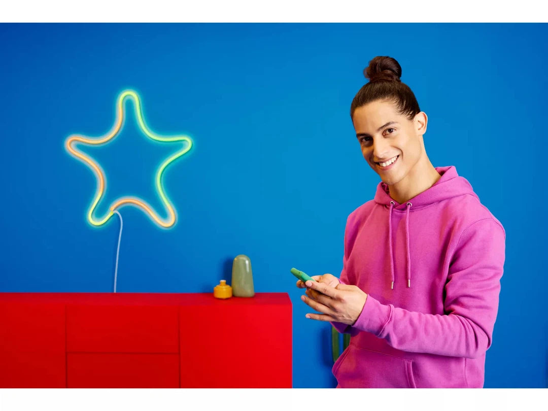 En person med langt hår i en knold står foran en blå væg og holder en smartphone i hånden iført en pink hættetrøje der smiler mens en farverig neon fleksibel lysstribe i form af en stjerne bliver vist i baggrunden grøn og orange farve dekorationer er på en rød bordplade
