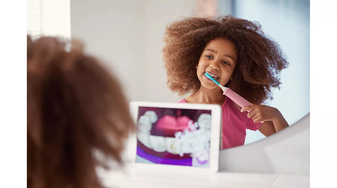 Pige med krøllet hår børster tænderne med en Philips Sonicare HX6352/42 For Kids sonisk eltandbørste foran et spejl, hvor der vises tandplejeillustrationer på en tablet