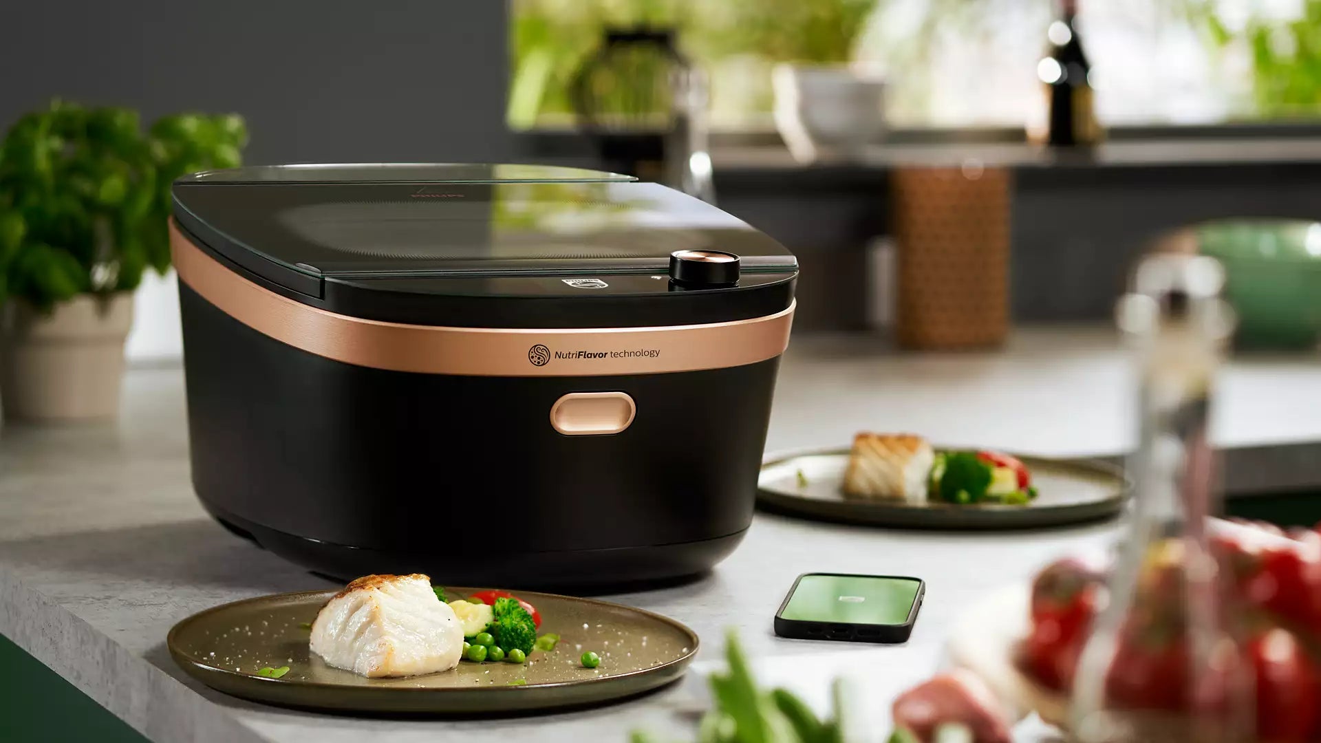 Philips NX0960/90 Air Steam Cooker i sort og kobberfarvet design placeret på en køkkenbordplade med dampede grøntsager og fisk på en tallerken ved siden af. En smartphone ligger på bordet og viser en app, der muligvis kontrollerer dampkogeren i en moderne køkkenindretning med planter i baggrunden.