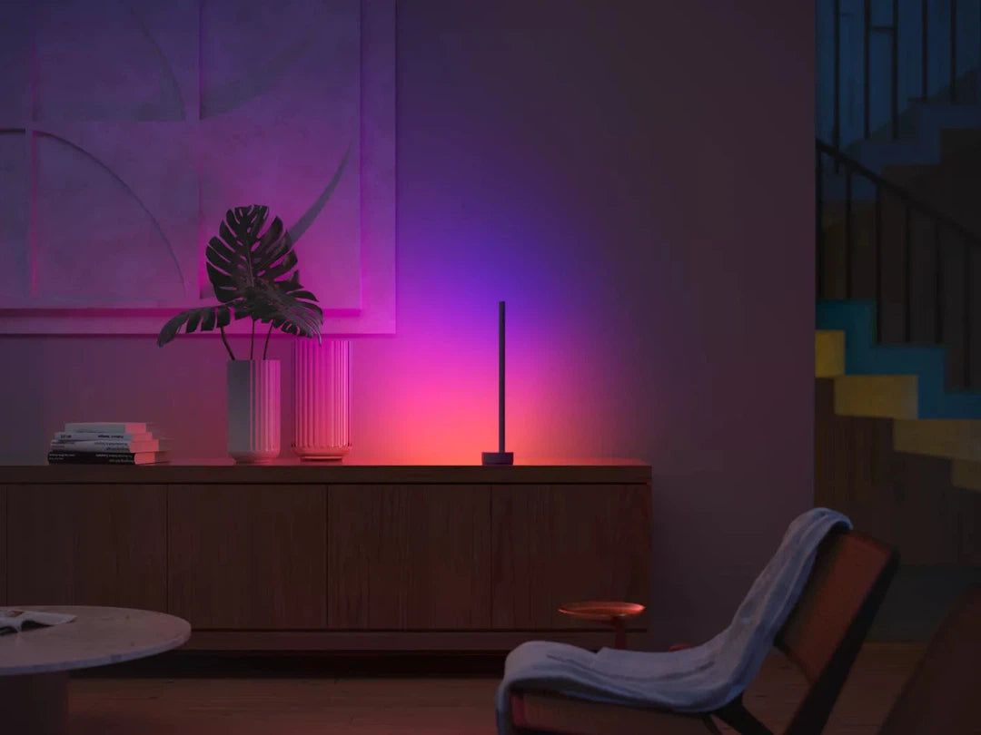 Philips Hue Signe gradient bordlampe i et moderne hjemmemiljø med bløde farver som pink og lilla der skaber en atmosfærisk belysning på et stilrent møbel med bøger og planter