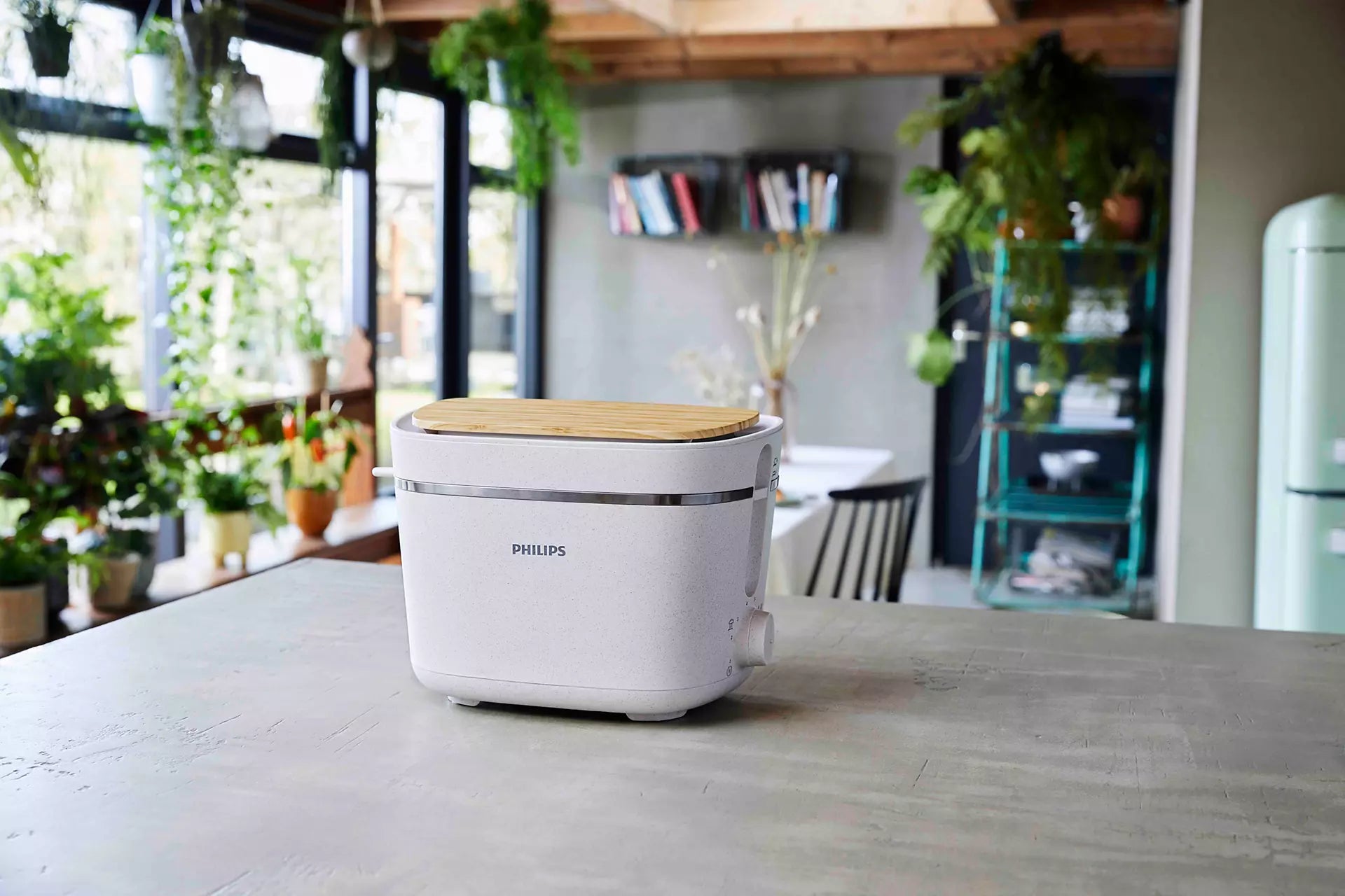 Philips HD2640/10 Eco Conscious Edition brødrister står på en grå bordplade i et moderne køkken med grønne planter i baggrunden og naturligt lys der strømmer ind gennem store vinduer. Brødristeren har et lysegråt design med en bambuslåg og en metalramme, der giver et stilrent udtryk.