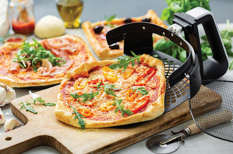 Philips HD9953/00 Airfryer XXL pizzabakke tilbehørssæt med en pizza på et skærebræt garnert med ruccola og peberfrugter foran flere pizzatyper og ingredienser som hvidløg og olivenolie i et lyst køkkenmiljø