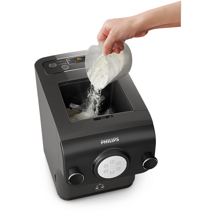 En person hælder mel fra en målebæger ind i en Philips HR2382/15 Avance Collection pastamaskine med digitalt display og knapper på toppen. Maskinen er sort og har en rund udskæringsplade på fronten.
