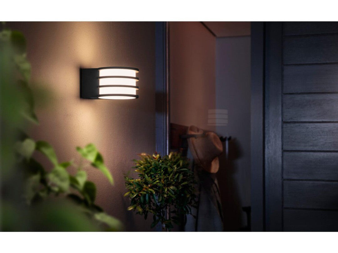 Philips HUE Lucca udendørs vægbelysning monteret på en væg med en moderne design og en blød lyseffekt omgivet af grønne planter