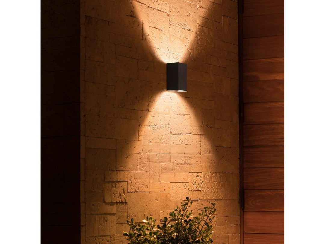 Philips Hue Resonate udendørs vægbelysning monteret på en væg af sten med et stilfuldt design der skaber en blød belysning og skygger i en udendørs indstilling med planter foran