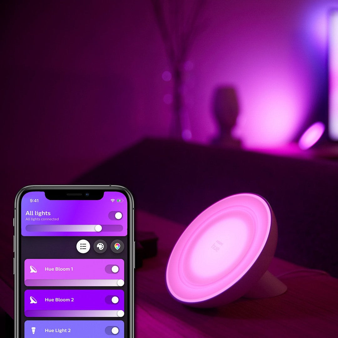 Philips Hue White and color ambiance Bloom bordlampe i en blød rosa farve placeret på et bord ved siden af en smartphone med Hue appen åben på skærmen i mørk baggrund
