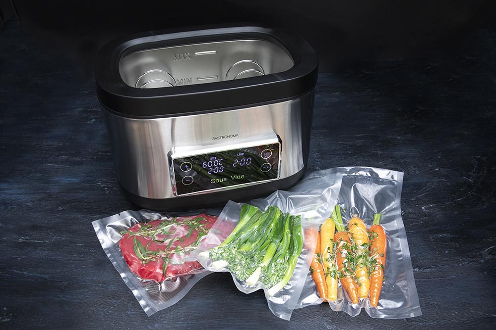 Billede af Gastronoma Sous Vide kar i sort og rustfrit stål med en kapacitet på 9 liter og en effekt på 700 watt, placeret ved siden af to vakuumforseglede poser med grøntsager og kød. Karret har en digital kontrolskærm med indstillinger for temperatur og tid. Grøntsagerne inkluderer gulerødder og grønne asparges, mens kødet er skåret i skiver og dekoreret med urter.