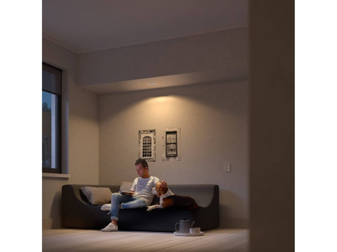 Philips HUE Milliskin indbygningsspot i hvid ambiance, installeret over en sofa, hvor en mand sidder og læser med en hund på skødet i et hyggeligt oplyst rum