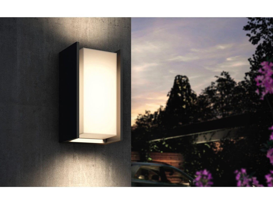 Philips HUE Turaco udendørs væglampe monteret på en grå væg med blød belysning i skumringen omgivet af grønne planter og blomster