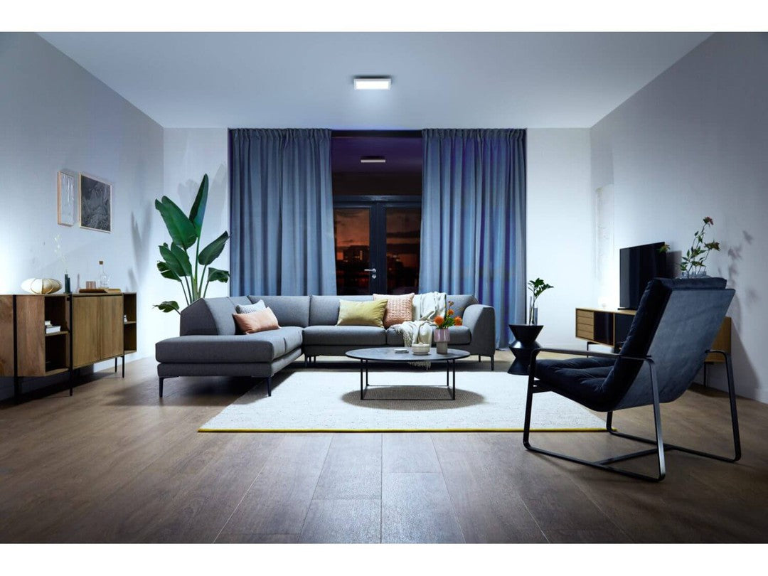 Indendørs stue med Philips HUE Aurelle loftpanel monteret i loftet, moderne sofa med puder, nøje udvalgte planter, et rundt bord med lys og dekorationer samt en tv-enhed, der giver en hyggelig og stilfuld atmosfære.