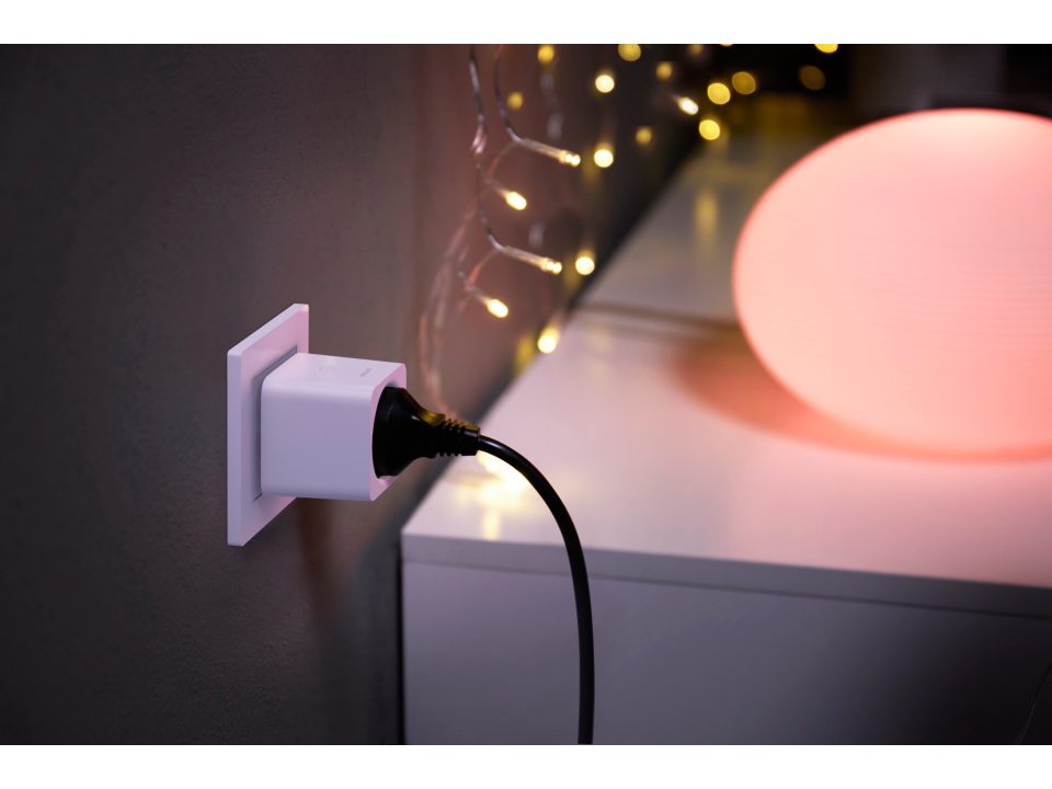 Philips Hue smart stikprop tilsluttet en vægkontakt med et sort kabel der strækker sig hen mod en lampe med blødt pink lys i baggrunden